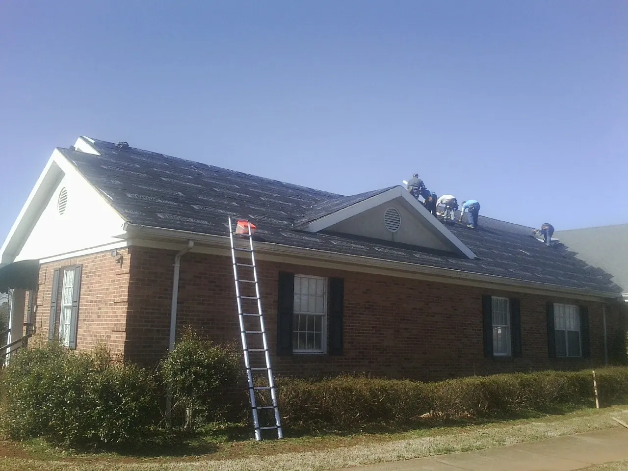 Crew tearing shingles off a brick ranch roof with a ladder set — Emergency Roof Repair work by our crew in South Royalton