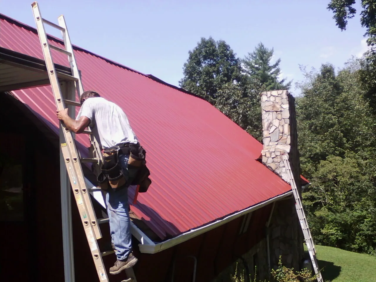 Roofer on a ladder beside a red metal roof and stone chimney — Soffit & Fascia Repair by Roofing Brothers in South Royalton